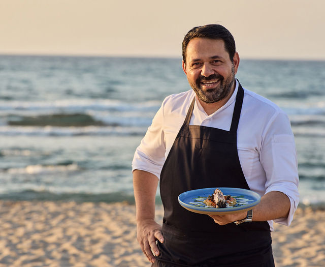 Paralos Kosta Alimia cook holding a gourmet dish with the sea in the background